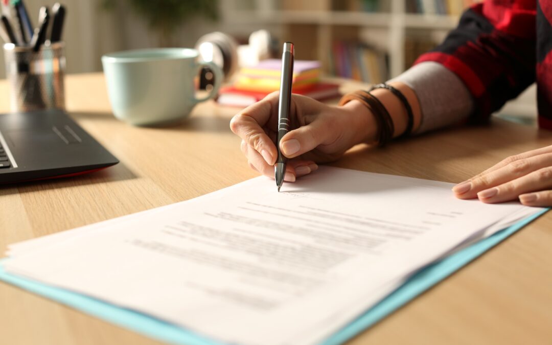 A woman sitting at a desk writing on paper next to a coffee mug and laptop.