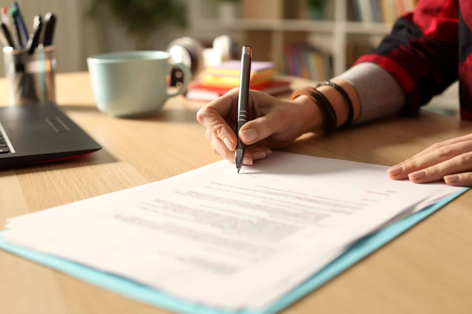 A woman sitting at a desk writing on paper next to a coffee mug and laptop.