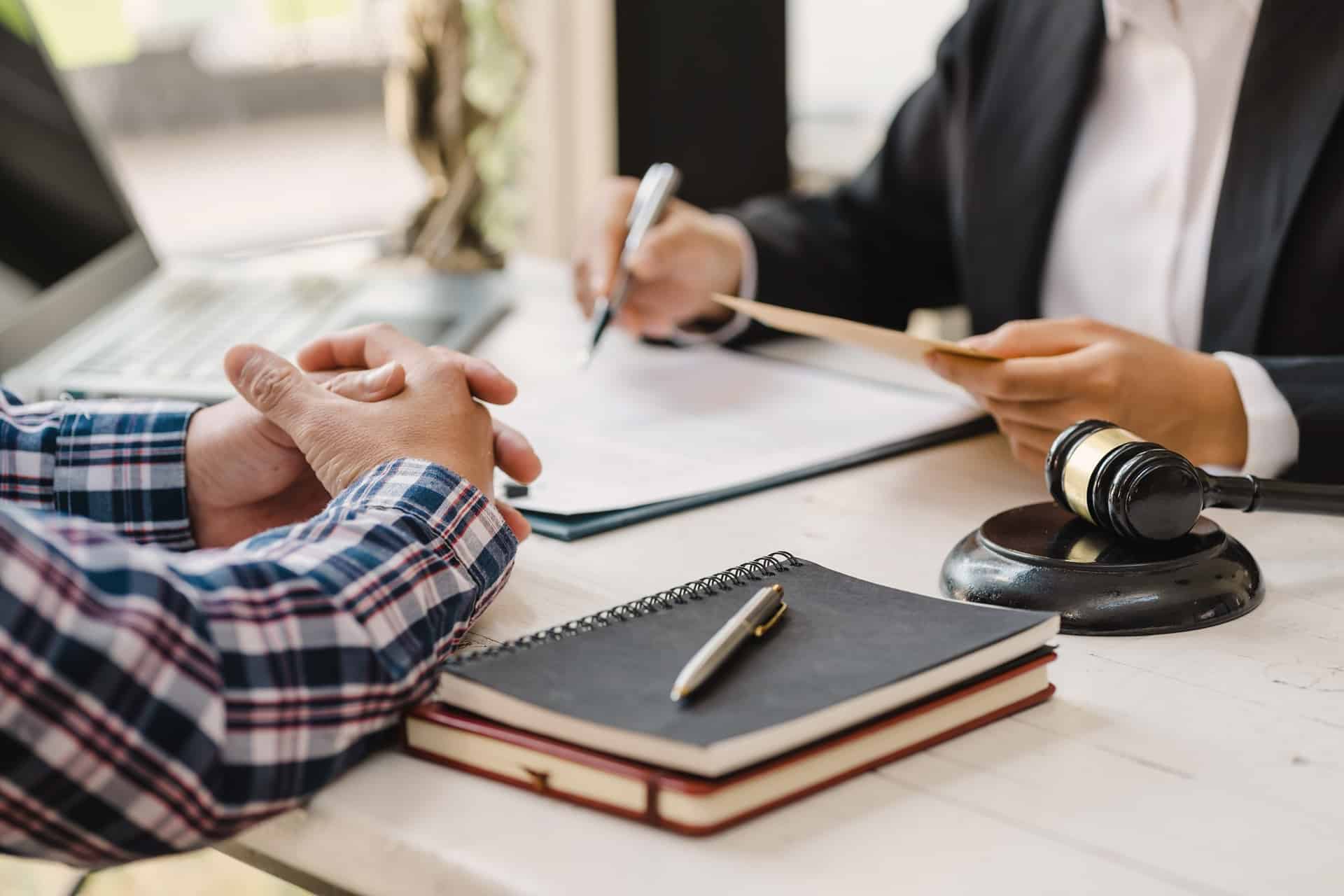 A female attorney holding a pen sitting at a desk across from a male client next to a notepad and gavel.