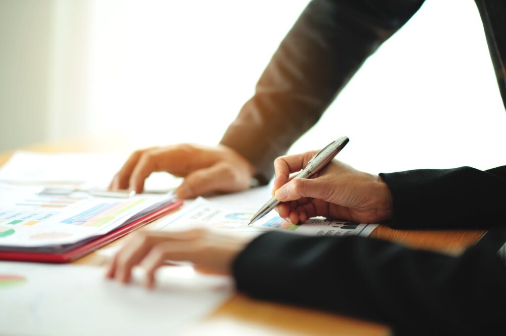 A person holding a pen writes on a piece of paper while a person standing next to them rests their hand on the table.