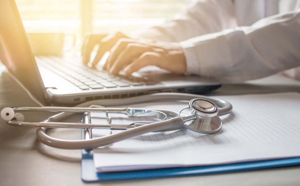 A person typing on a silver laptop at a desk next to a clipboard with a stethoscope resting on it.
