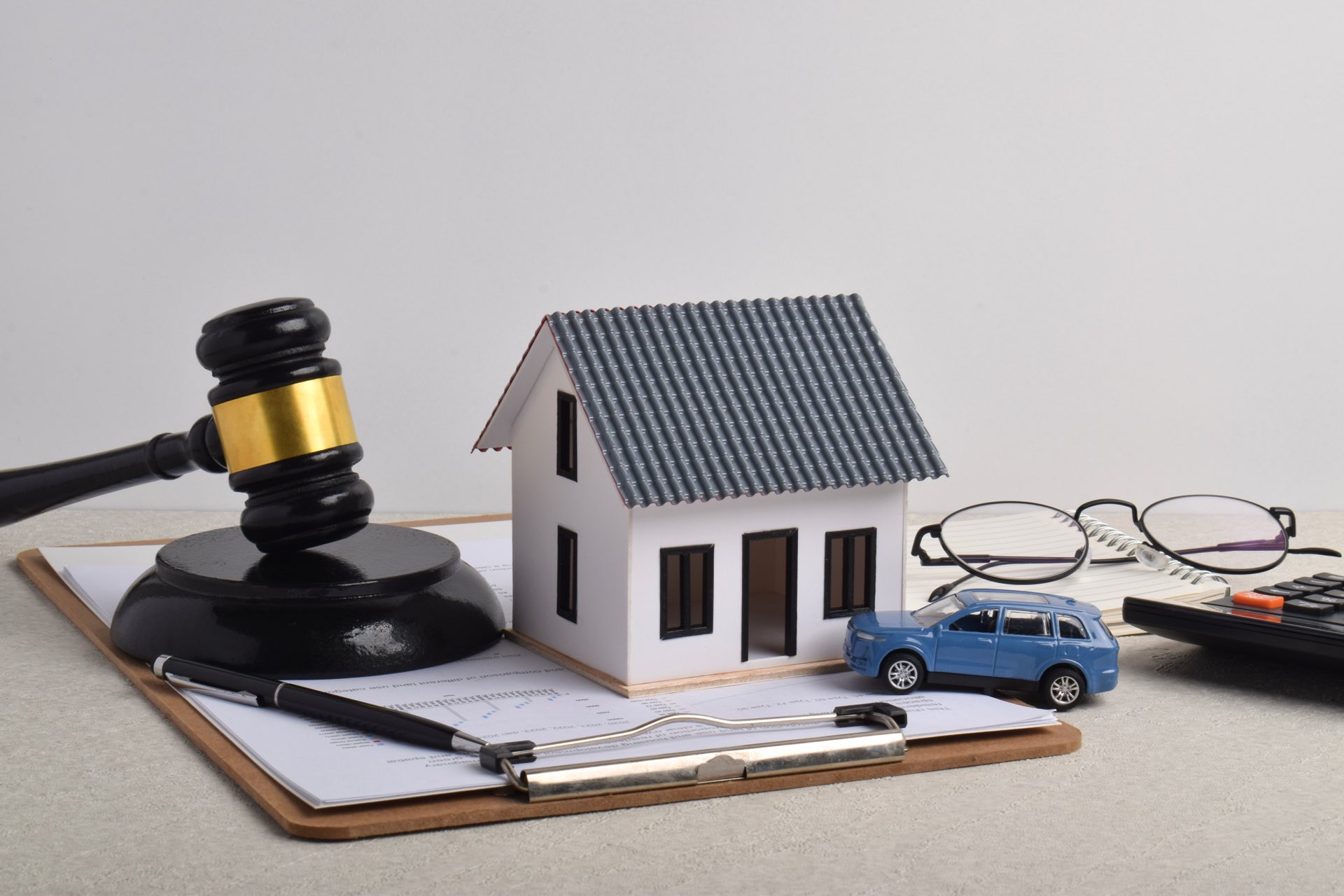 A gavel, toy house and car, glasses, calculator on top of a clipboard of papers