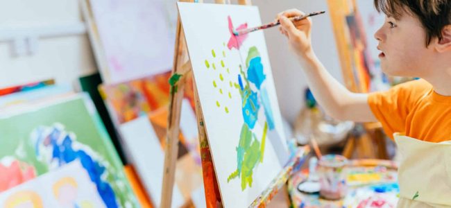 Close up shot of caucasian boy with Down's syndrome painting with easel in art class at school. Concept disabled kid learning.